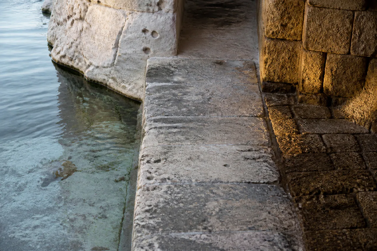 Continuous pale stone threshold with cool still seawater on one side and late sun held in darker masonry on the other